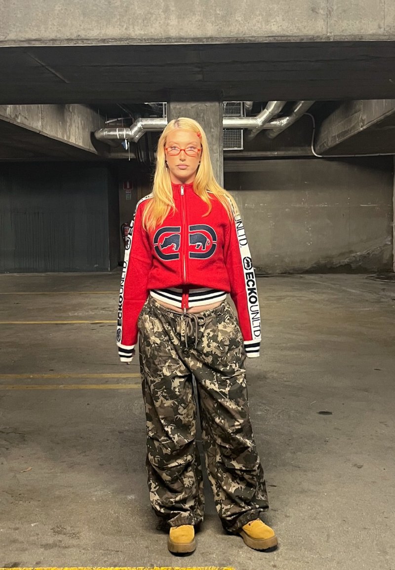 Piros cipzáras dzseki fekete és fehér kiegészítőkkel, katonai stílusú, bő nadrág, és homokszínű lábbeli. Figyelemre méltó kerek szemüveg és hosszú szőke haj.