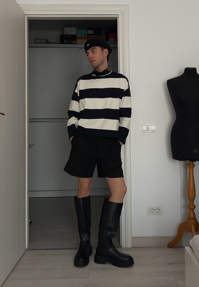 Botas negras hasta la rodilla, un suéter a rayas azul marino y blanco, y pantalones cortos negros están en una habitación con suelo de madera clara y un maniquí cercano.