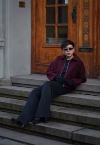 Giacca borgogna strutturata sopra una maglietta nera, abbinata a pantaloni larghi a righe verticali neri. Guanti scuri e occhiali da sole completano il look.