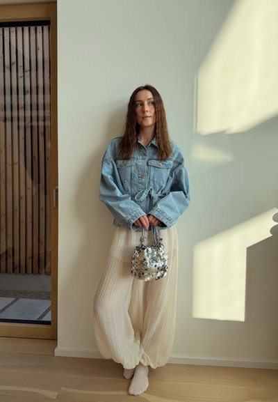 Mujer con el cabello largo y castaño, vestida con una chaqueta vaquera grande, pantalones sueltos color crema, sosteniendo un bolso plateado de lentejuelas, de pie descalza junto a una pared iluminada por el sol.
