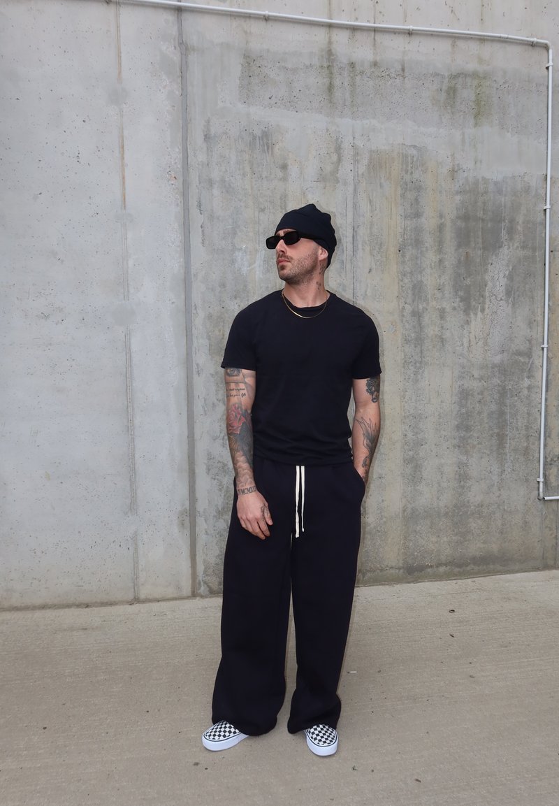 Man met tatoeages die een zwarte muts, zonnebril, T-shirt, wijde broek en geruite instappers draagt, staand tegen een betonnen muur.