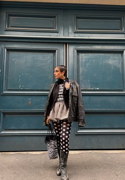 Chaqueta oversized de cuero negro, camiseta a cuadros, medias con lunares, botas vaqueras plateadas, bolsa de estampado de cebra con borde de pelo, frente a unas puertas azules.