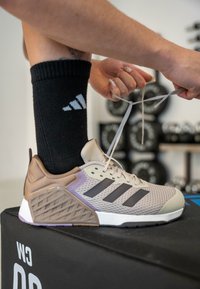 Szürke és bézs atlétikai cipő lila kiemelésekkel. Hálós felsőrésze, három fekete csíkja és texturált sarka van. A fűzőket éppen megkötik.