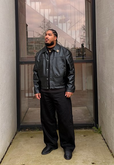 Hombre con cabello trenzado que lleva una chaqueta de cuero negra, pantalones negros y zapatos negros, de pie entre paredes de hormigón frente a una puerta de vidrio.