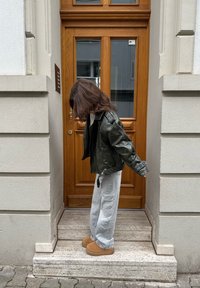 Zielona skórzana kurtka, szare luźne dżinsy, beżowe buty na platformie; kobieta stojąca na schodach obok drewnianych drzwi z szybami.