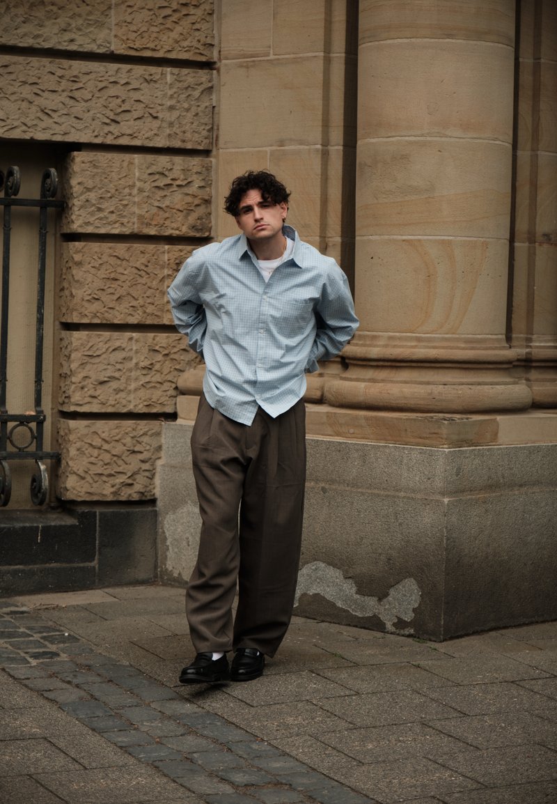 Νέος άνδρας με μπλε καρό πουκάμισο και καφέ παντελόνι στέκεται σε αστικό πεζοδρόμιο, γέρνοντας ελαφρώς με τα χέρια πίσω από την πλάτη κοντά σε πέτρινο κτίριο.