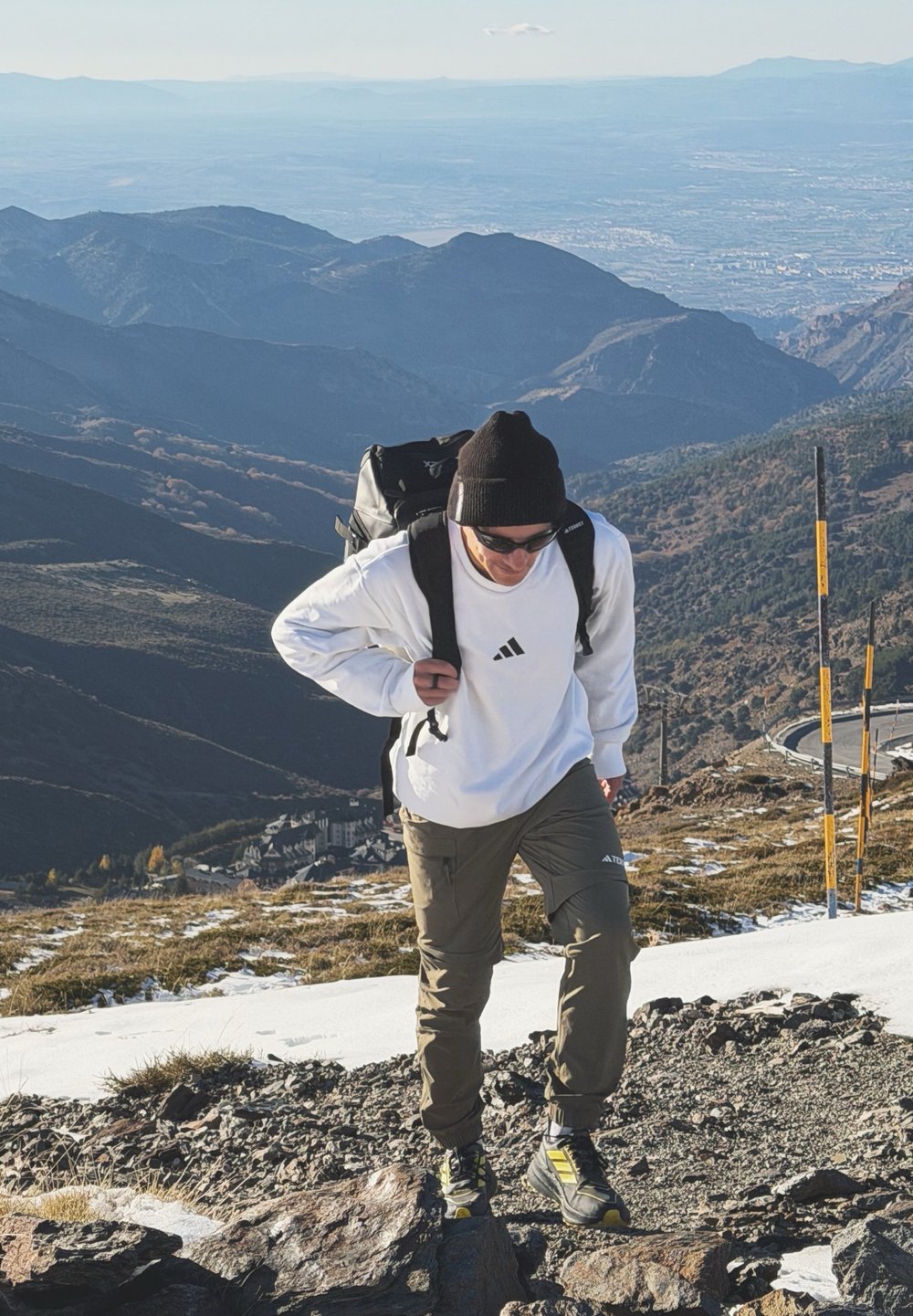 Homme portant des lunettes de soleil, un bonnet et un sac à dos, grimpant une pente raide sur un sentier montagneux rocailleux et enneigé, avec une chaîne de montagnes au loin et une vallée en contrebas.