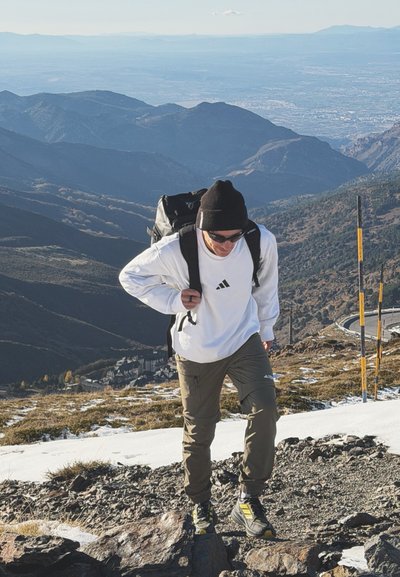 Hombre con gafas de sol, gorro y mochila caminando cuesta arriba por un sendero rocoso y nevado en la montaña, con una cadena montañosa y un valle a lo lejos.