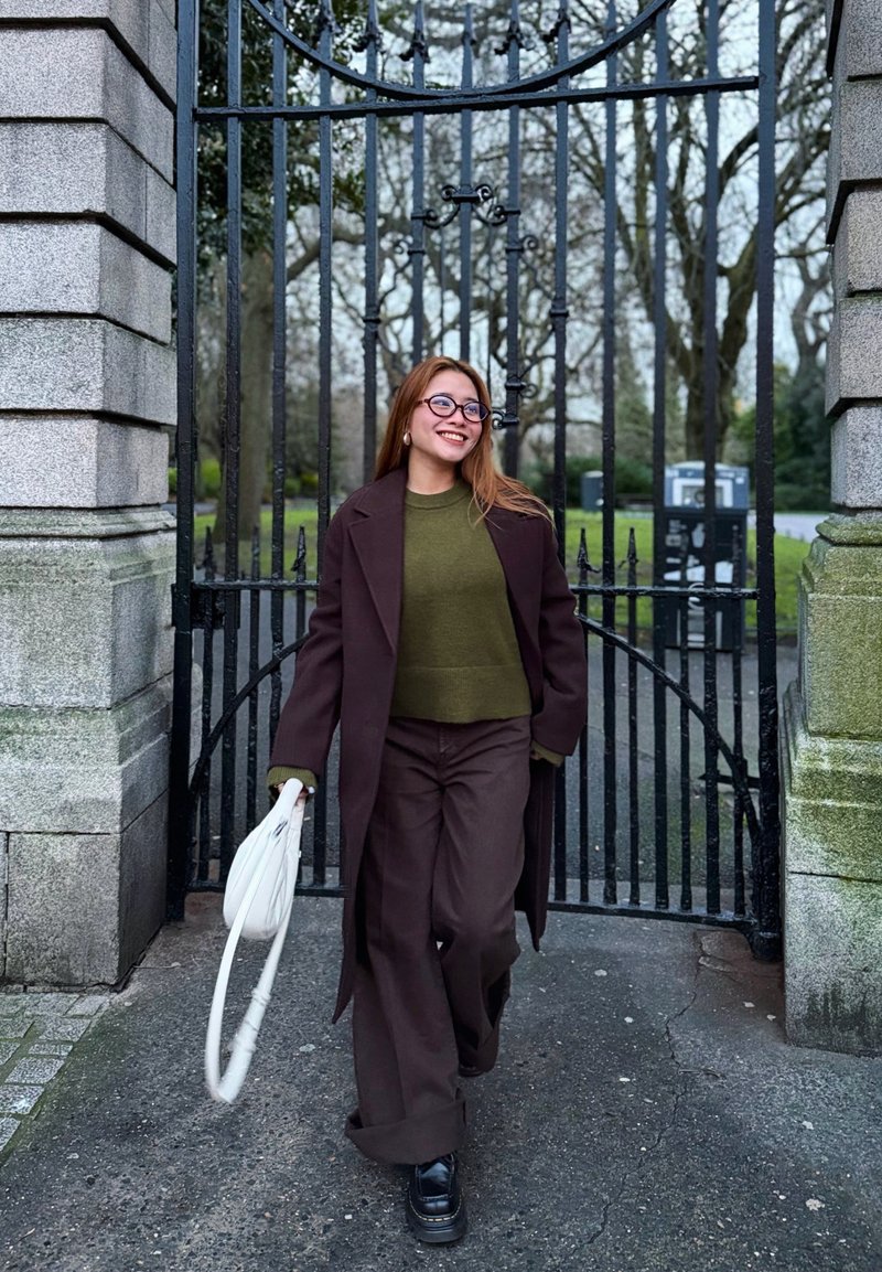 Jeune femme souriante marchant à travers une grille en fer noir, portant un manteau et un pantalon marron, un pull kaki, des chaussures noires et tenant un sac blanc.