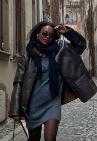 Hnědá kožená oversized bunda, šedé šaty, černý šál a sluneční brýle. Kamenitá ulice se starobylými budovami a břečťanem.