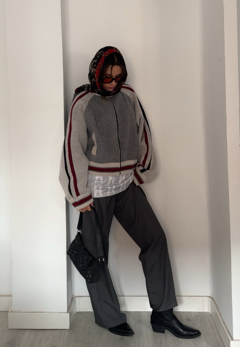 Cardigan oversized em cinza e vermelho, com fecho de correr e punhos listrados, usado sobre uma parte de cima branca acolchoada, combinado com calças escuras e botas de tornozelo pretas.