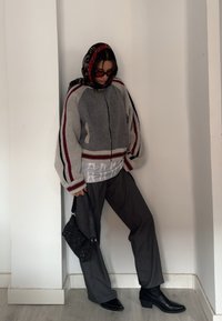Cardigan oversized em cinza e vermelho, com fecho de correr e punhos listrados, usado sobre uma parte de cima branca acolchoada, combinado com calças escuras e botas de tornozelo pretas.