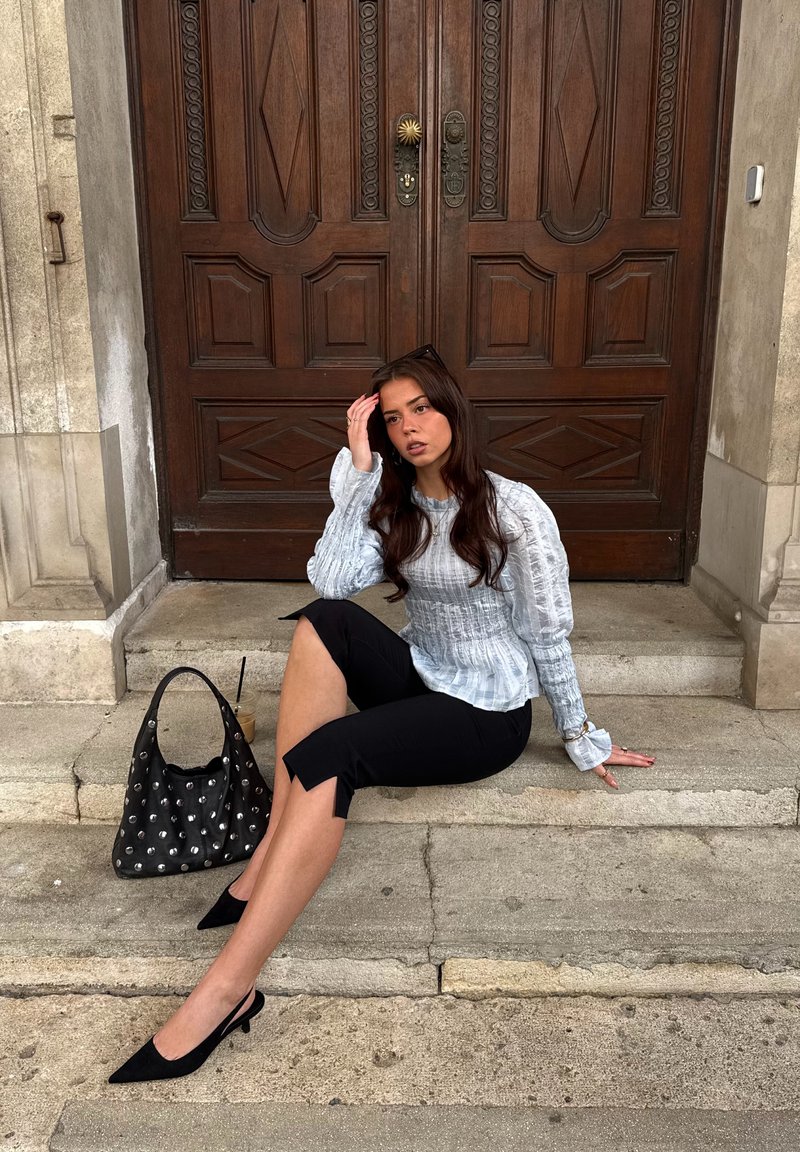 Νεαρή γυναίκα με μακριά σκούρα μαλλιά, φορώντας λευκό μπλουζάκι και μαύρο παντελόνι, καθισμένη σε πέτρινα σκαλοπάτια μπροστά από μεγάλες ξύλινες πόρτες, δίπλα σε μια μαύρη τσάντα με καρφιά.