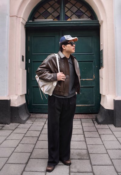 Persona que lleva una chaqueta de cuero marrón, suéter gris, pantalones negros, gorra y gafas de sol, llevando una bolsa blanca, de pie en una calle empedrada.