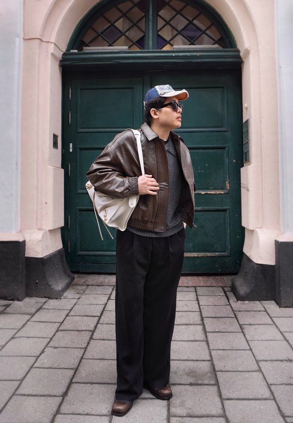 Person som har på seg en brun skinnjakke, grå genser, svarte bukser, caps og solbriller, med en hvit veske, står på en brosteinsgate.