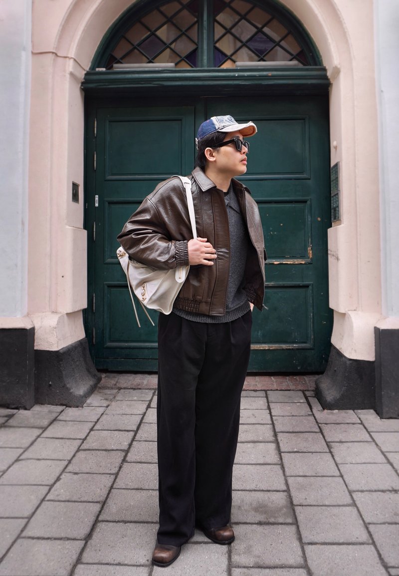 Personne portant une veste en cuir marron, un pull gris, un pantalon noir, une casquette et des lunettes de soleil, portant un sac blanc, debout dans une rue pavée.