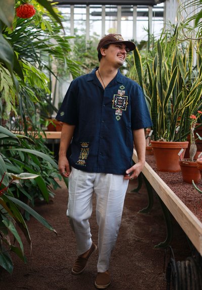 Hombre con camisa azul marino estampada y pantalones blancos caminando y sonriendo en un invernadero rodeado de varias plantas en macetas.