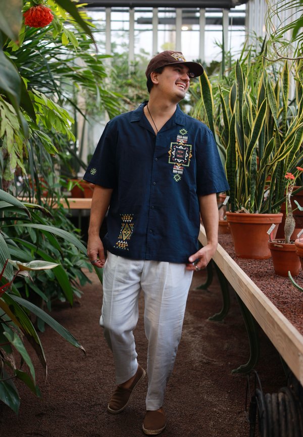 Mees tumesinise mustriga särgis ja valgetes pükstes kõnnib ja naeratab kasvuhoones, mida ümbritsevad erinevad potitaimed.