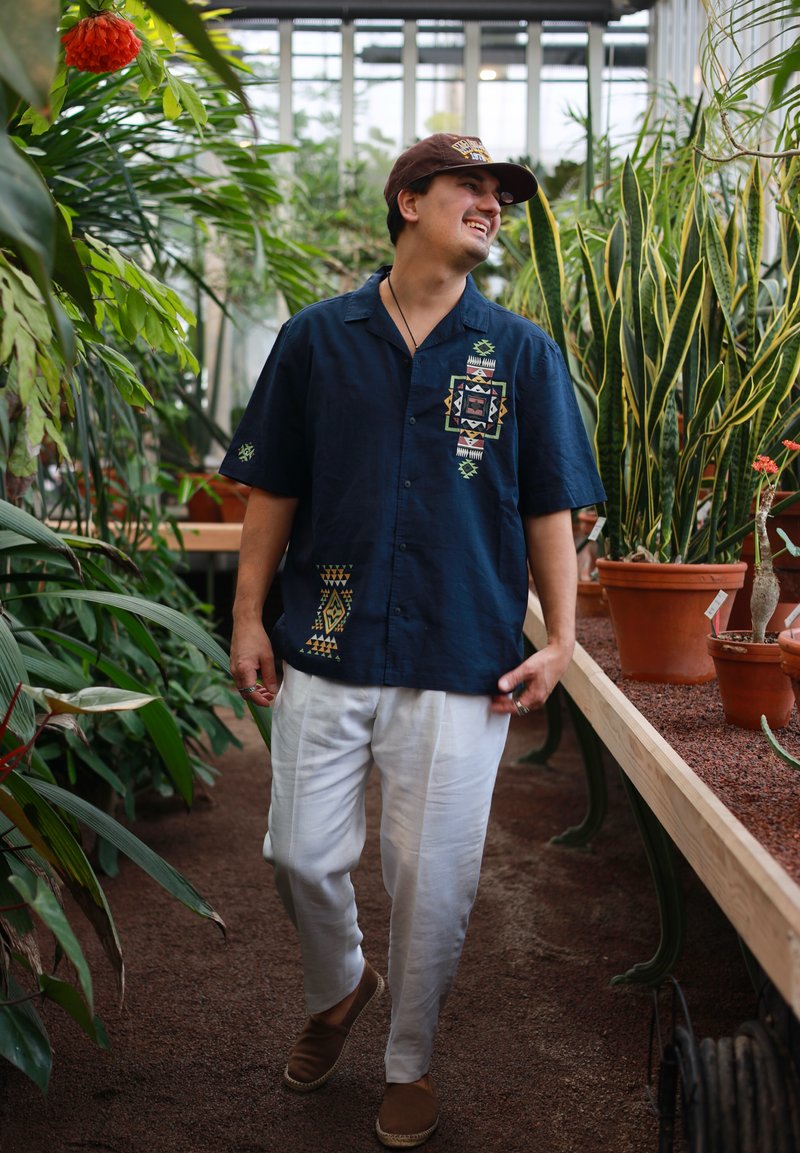 Mann in navyblauem, gemustertem Hemd und weißer Hose, der in einem Gewächshaus spazieren geht und lächelt, umgeben von verschiedenen Topfpflanzen.