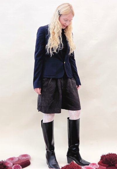 Botas de cuero negras hasta la rodilla, medias blancas texturizadas, falda oscura y americana azul marino; el modelo está de pie contra un fondo claro, con alfombras rosas y rojas cerca.