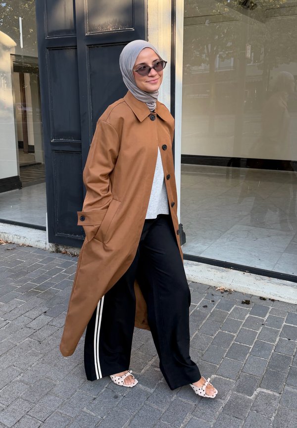 Vrouw met een grijze hijab, een lange bruine jas, zwarte broek met witte strepen, zonnebril en witte sandalen met stippen, lopend op het trottoir.