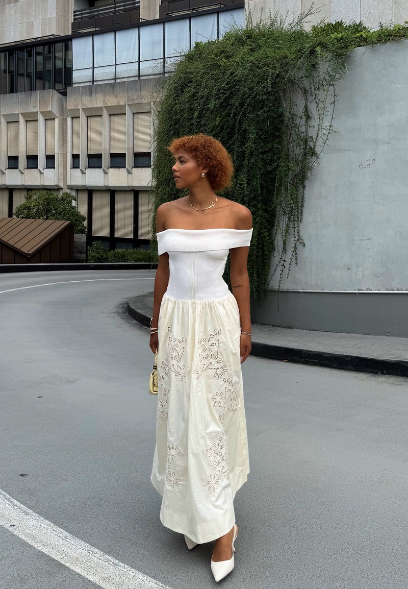 Donna con capelli ricci che indossa un vestito bianco a spalle scoperte con ricami floreali e tacchi bianchi, in piedi su una strada urbana accanto a zone verdi.