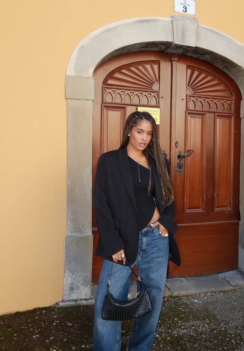 Blazer preto oversize, top preto justo, jeans azuis soltos e uma pequena mala preta trançada. Fundo com uma porta de madeira com detalhes ornamentados.