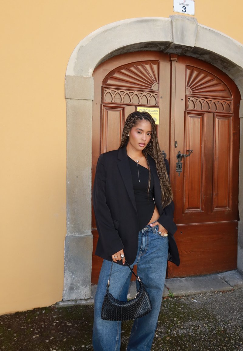 Svart överdimensionerad kavaj, tajt svart topp, lösa blå jeans av denim och en liten svart vävd handväska. Bakgrunden visar en trädörr med utsmyckade detaljer.