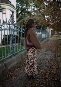 Woman wearing a brown fleece jacket and leopard print skirt stands on a leaf-covered sidewalk beside a decorative metal fence and autumn trees.