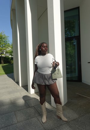 Top blanco texturizado, pantalones cortos a cuadros en blanco y negro, botas de vaquero beige y un bolso verde claro. De pie junto a columnas blancas y vegetación.