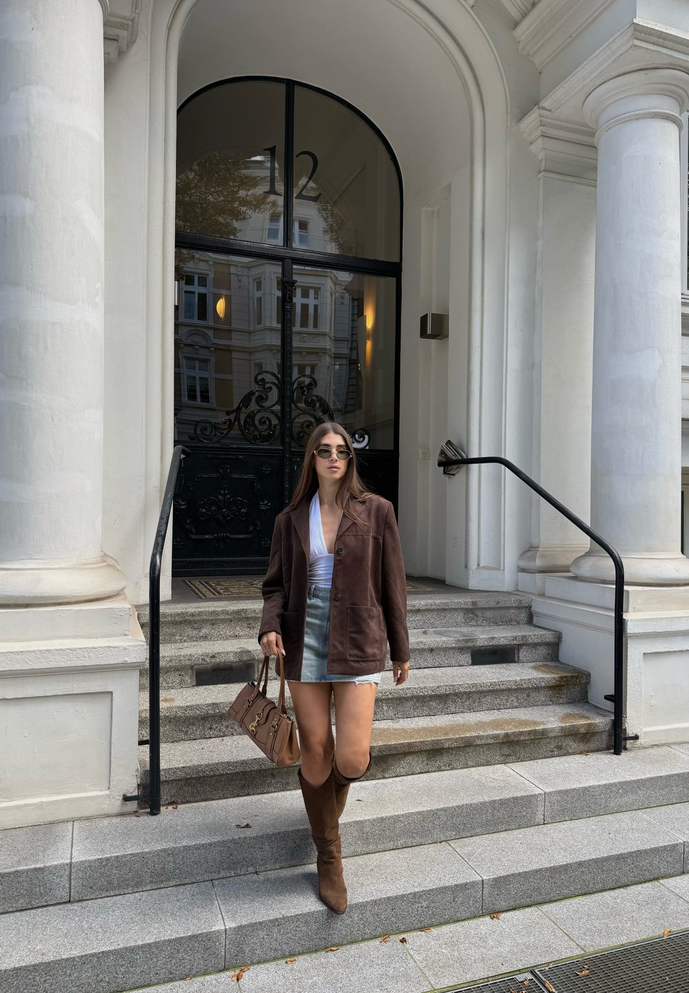 Chaqueta de ante marrón, blusa blanca, falda de mezclilla; botas marrones hasta la rodilla. Sosteniendo un bolso beige. De pie en escalones de piedra frente a una puerta de vidrio.