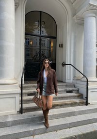 Chaqueta de ante marrón, blusa blanca, falda de mezclilla; botas marrones hasta la rodilla. Sosteniendo un bolso beige. De pie en escalones de piedra frente a una puerta de vidrio.