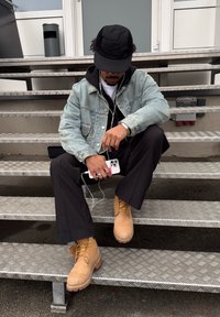 Homme assis sur des escaliers en métal, portant une veste en jean, une casquette noire, un pantalon noir, des bottes marron clair, tenant un smartphone avec des écouteurs.