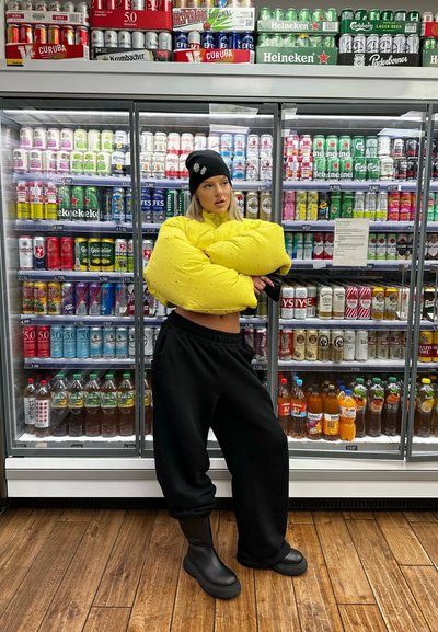 Chaqueta acolchada amarilla con superficie texturizada, pantalones deportivos negros y botines negros. El fondo presenta varias latas de bebida coloridas.