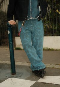 Jean bleu clair à motif avec une coupe ample, présentant un design abstrait subtil. Porté avec une veste sombre et des chaussures noires. Porte-clés attaché.