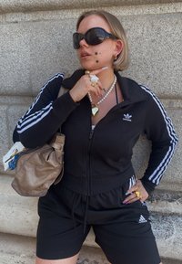 Sort ribbet zip-jakke med tre hvide striber, sorte shorts og en brun håndtaske. Tilbehør inkluderer lagdelte halskæder og neglekunst.