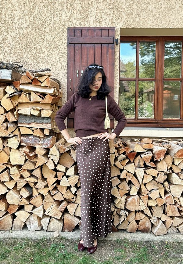 Femme aux cheveux foncés portant un pull marron, une jupe à pois et des lunettes de soleil, debout les mains sur les hanches à côté d'un tas de bois empilé devant une maison.