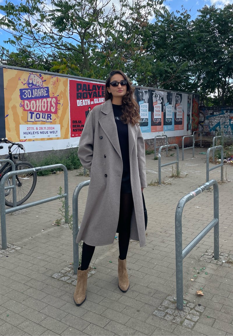Manteau en laine gris clair, à double boutonnage, avec de grands boutons noirs. Pull noir, leggings ajustés et bottines beige. Environnement extérieur avec des affiches.
