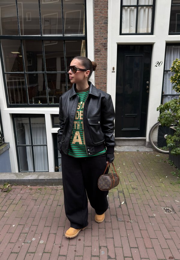 Žena u crnoj kožnoj jakni, zelenoj prugastoj majici, crnim hlačama, bež čizmama i sunčanim naočalama drži okruglu smeđu torbu dok hoda po ulici od cigle.