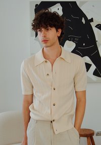 Camisa de botão em malha beige com textura canelada e mangas curtas; apresenta botões escuros e um colarinho clássico. Fundo claro.