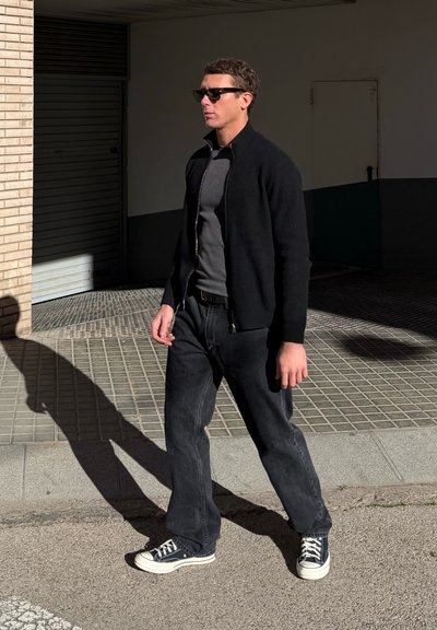 Hombre con gafas de sol negras, chaqueta negra con cremallera, camiseta gris, vaqueros negros holgados y zapatillas negras caminando por una acera iluminada por el sol.