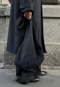 Άτομο με μαύρο παλτό και παντελόνι κρατάει μεγάλη μαύρη υφασμάτινη τσάντα ώμου, στέκεται σε αστικό πεζοδρόμιο μπροστά σε τοίχο με υφή από πέτρα.