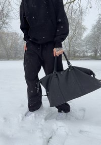 Sort skinn duffelbag med strukturert form og doble håndtak, plassert i snødekt utendørs omgivelser, brukt sammen med svart hettegenser og bukser.