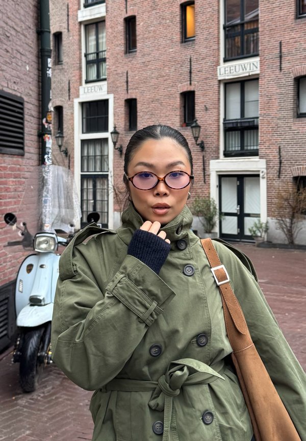 Osoba w zielonym płaszczu przeciwdeszczowym i okularach z fioletowymi szkłami stoi przed budynkiem z cegły, obok stoi zaparkowany jasnoniebieski skuter.