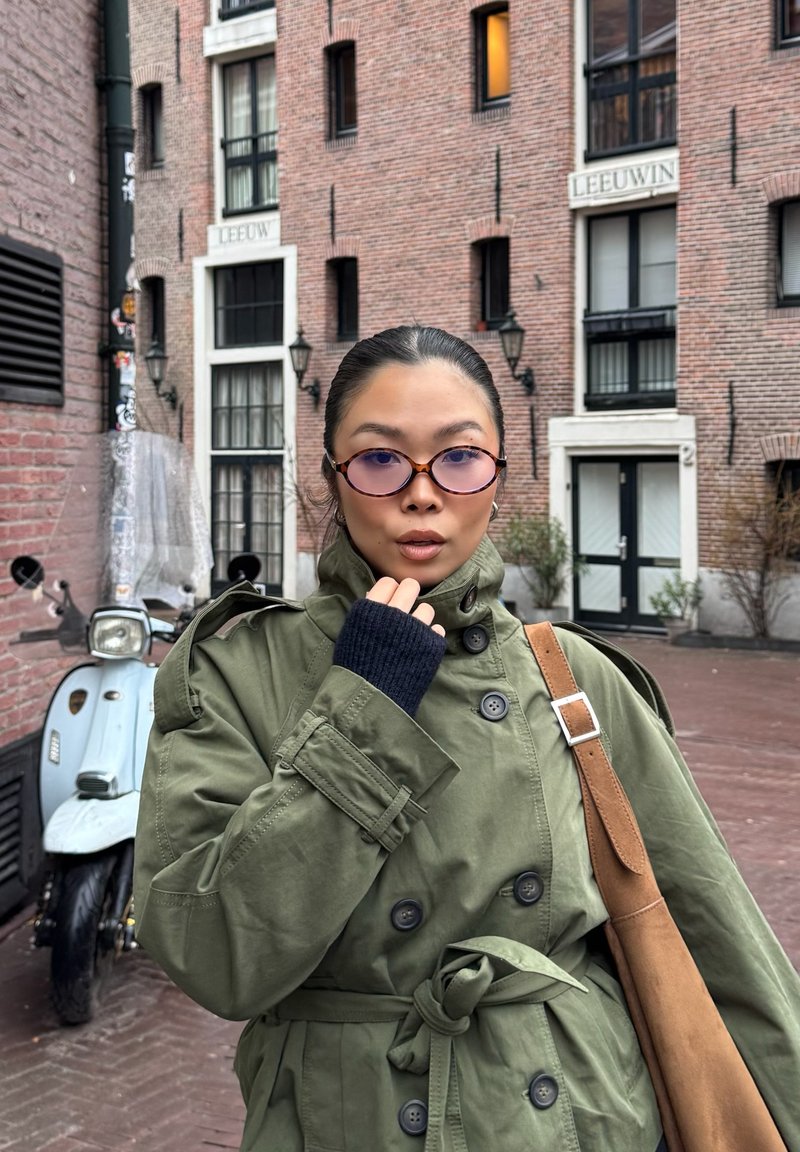 Personne portant un trench-coat vert et des lunettes teintées de violet se tient devant un bâtiment en briques avec un scooter bleu clair garé à proximité.