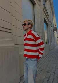 Sarkani un baltā krāsā svītraina garām piedurknēm polo krekls, brīva piegriezuma, valkāts kopā ar gaiši ziliem, vaļīgiem džinsiem, pilsētas vidē ar gaišu fonu.