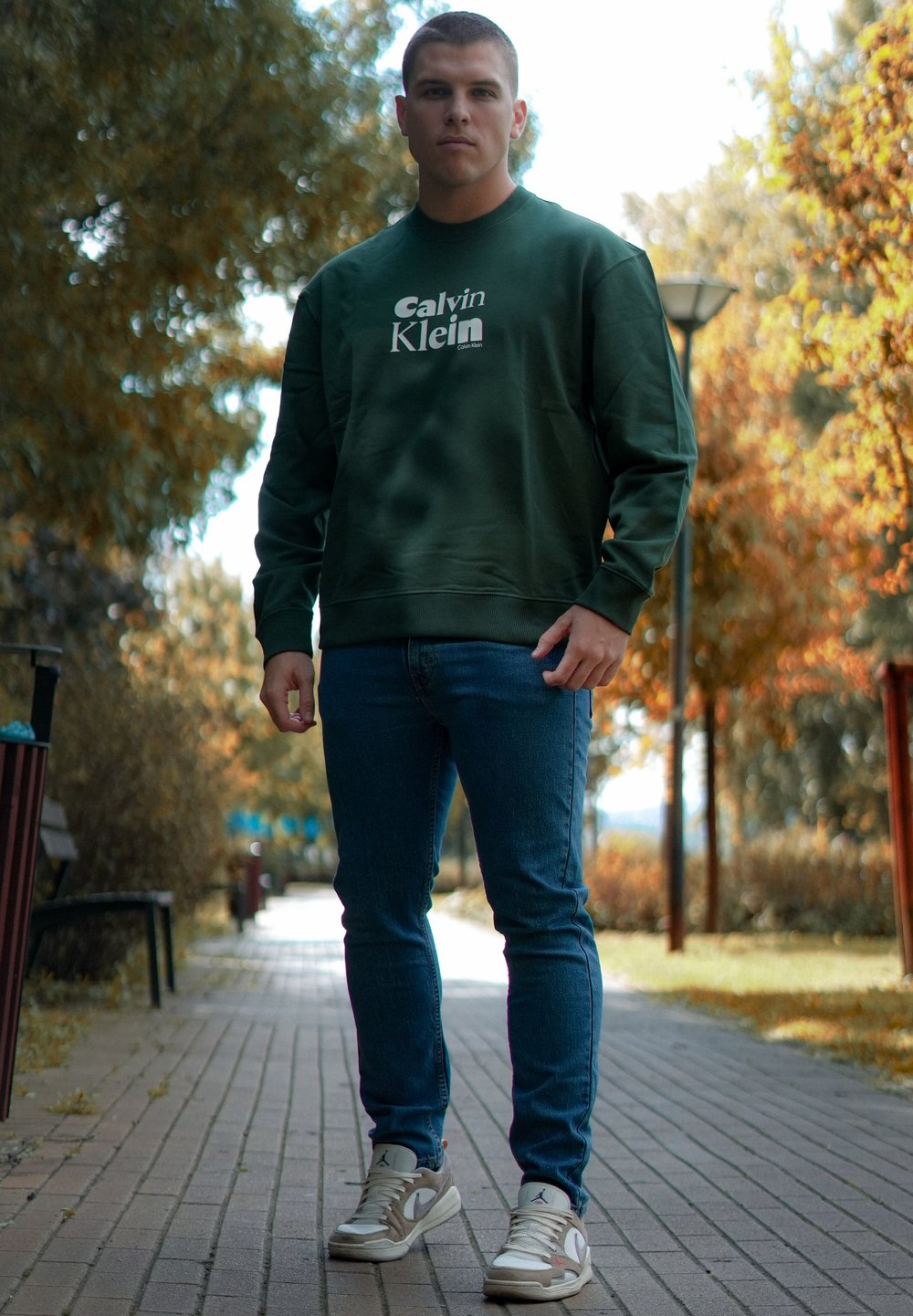 Grønn hettegenser med hvit logo, blå slank-fitt jeans og beige sneaks. Bakgrunnen har trær og en asfaltert sti.