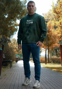 Grønn hettegenser med hvit logo, blå slank-fitt jeans og beige sneaks. Bakgrunnen har trær og en asfaltert sti.