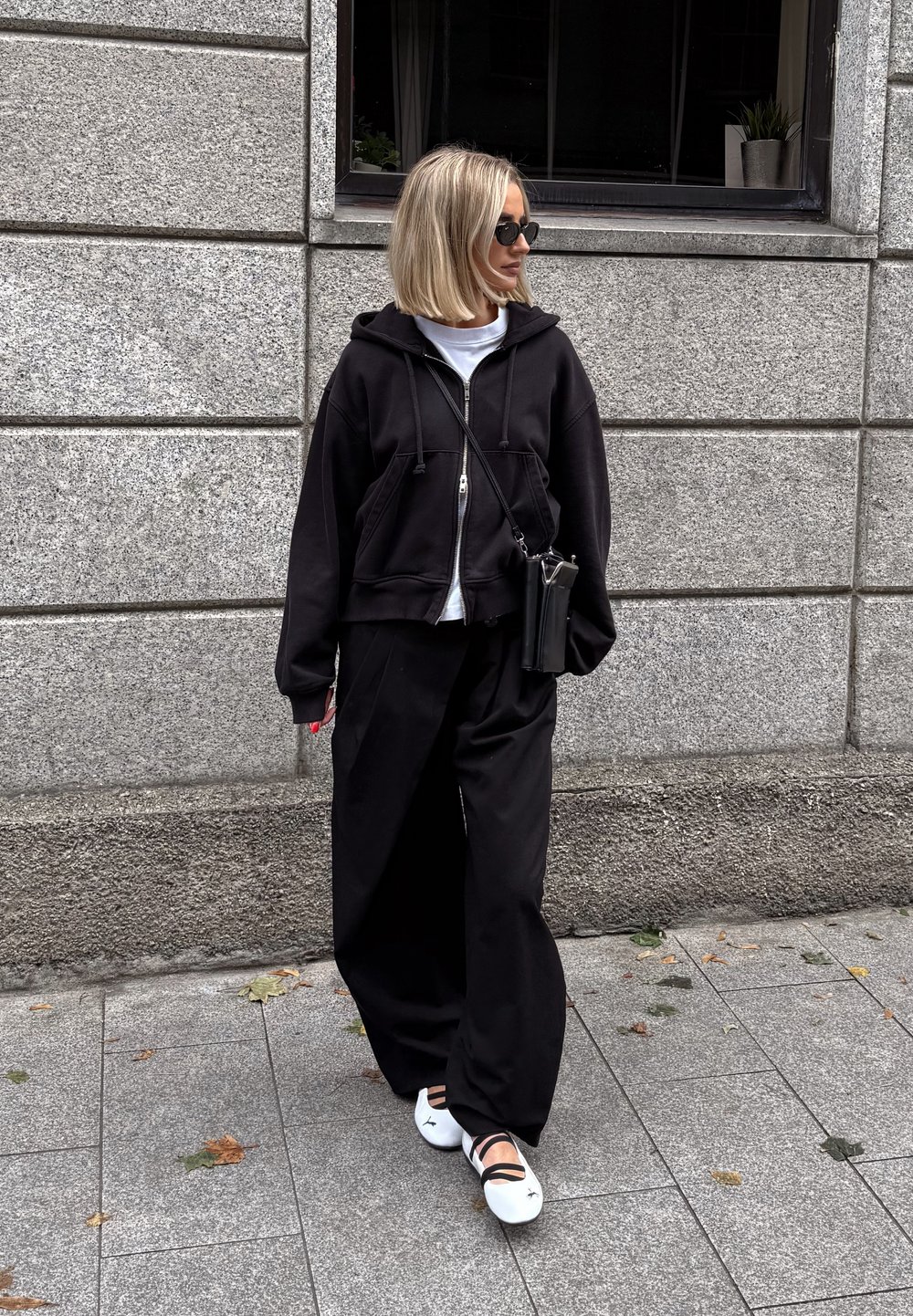 Felpa nera con zip sopra una maglietta bianca, pantaloni neri larghi e scarpe bianche con fasce nere. Accessori includono una borsa a tracolla nera.