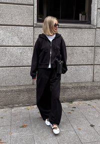 Felpa nera con zip sopra una maglietta bianca, pantaloni neri larghi e scarpe bianche con fasce nere. Accessori includono una borsa a tracolla nera.